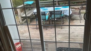 Une vue à travers une porte vitrée d’une terrasse en bois, avec des meubles d’extérieur et une piscine.