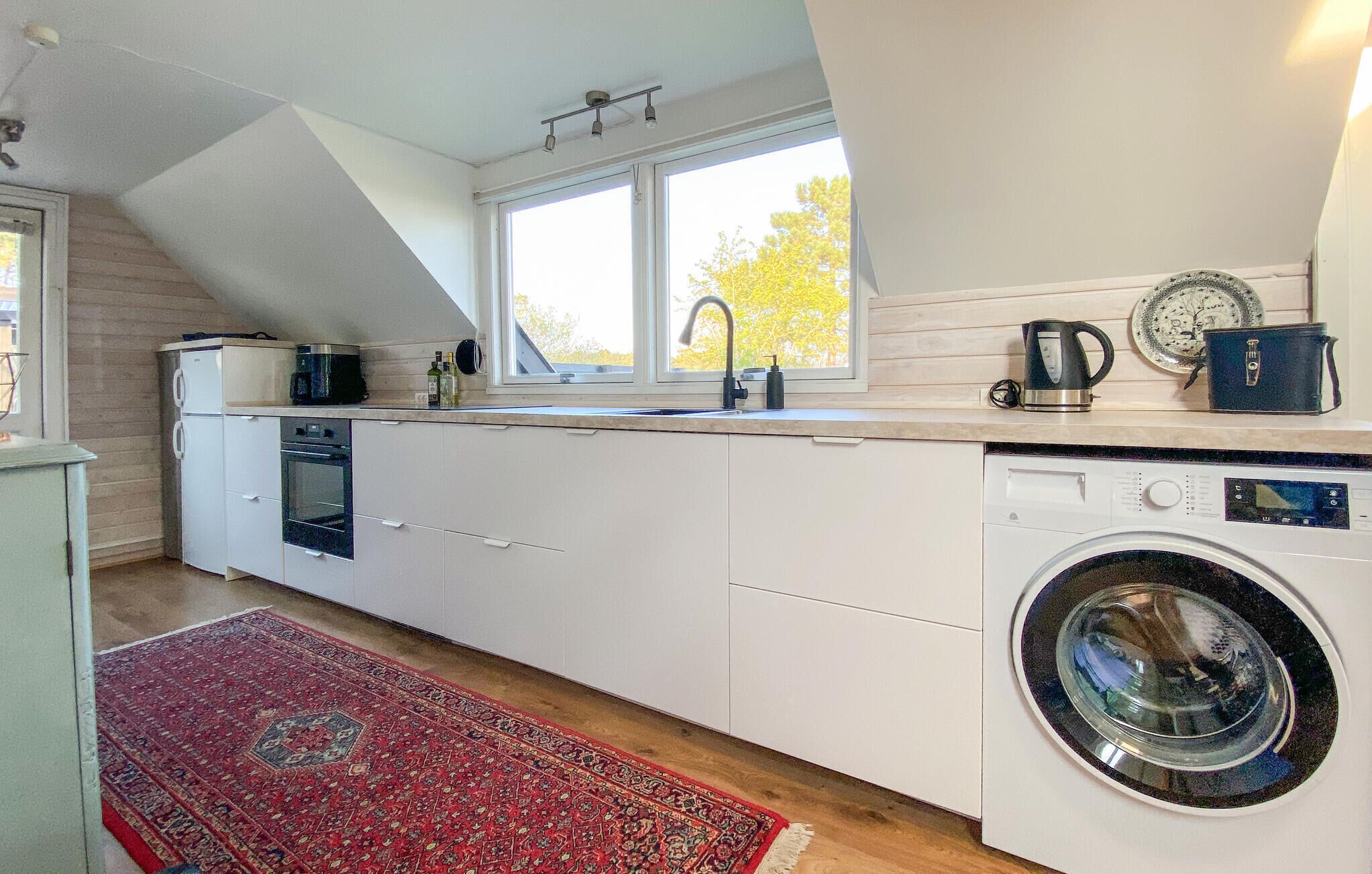 Une cuisine compacte équipée d’un lave-linge, d’une bouilloire et d’une fenêtre donnant sur les arbres.