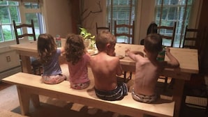 Quatre enfants sont assis autour d’une table en bois dans une pièce dotée de grandes fenêtres et d’une plante.