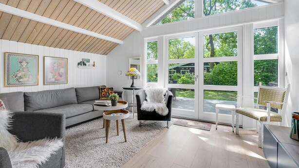 Een ruime kamer met een hoge plafond, grote ramen, een grijze bank, een witte pluizige fauteuil en een klein tafeltje met een lamp.