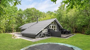 Een moderne cabin met een schuin dak, een terras met zitplaatsen buiten, en een trampolaan op de voorgrond.