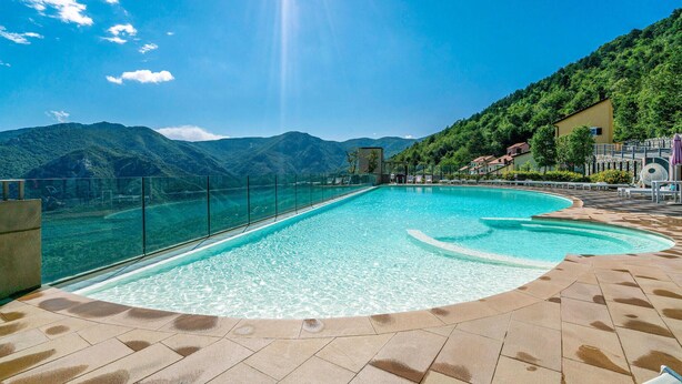 Une piscine d’un bleu éclatant, séparée du reste par une barrière de verre, entourée d’une terrasse carrelée, le tout se fondant dans un paysage de montagnes et d’un ciel dégagé.
