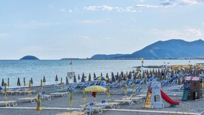 Une plage bordée de rangées de chaises longues et de parasols, une aire de jeux et une montagne en arrière-plan.