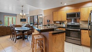 Una cocina moderna con gabinetes de madera, electrodomésticos de acero inoxidable, una zona de comedor con una mesa redonda y sillas, y un amplio ventanal que proporciona luz natural.