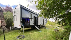 A recreational vehicle with a sliding door, a small outdoor area with steps, and a hose on the ground. - 2022 5th Wheel RV, 2BD, Sleeps 6, Kootenai River Views/Access, Picnic Table (Bonners Ferry)