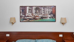 Une chambre d’hôtel avec un lit, deux lampes fixées au mur et un tableau encadré représentant un bâtiment historique avec une fontaine.