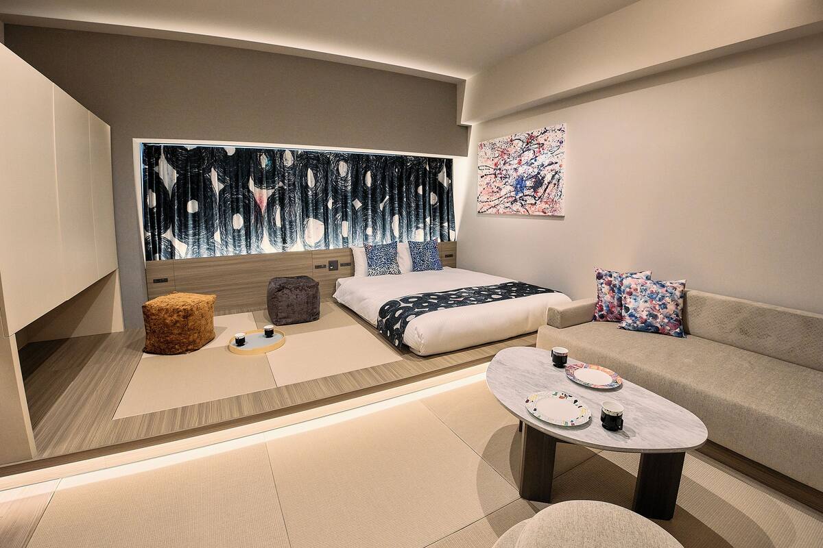 A modern, well-lit bedroom with a large bed, a sofa, a coffee table, and a small ottoman.