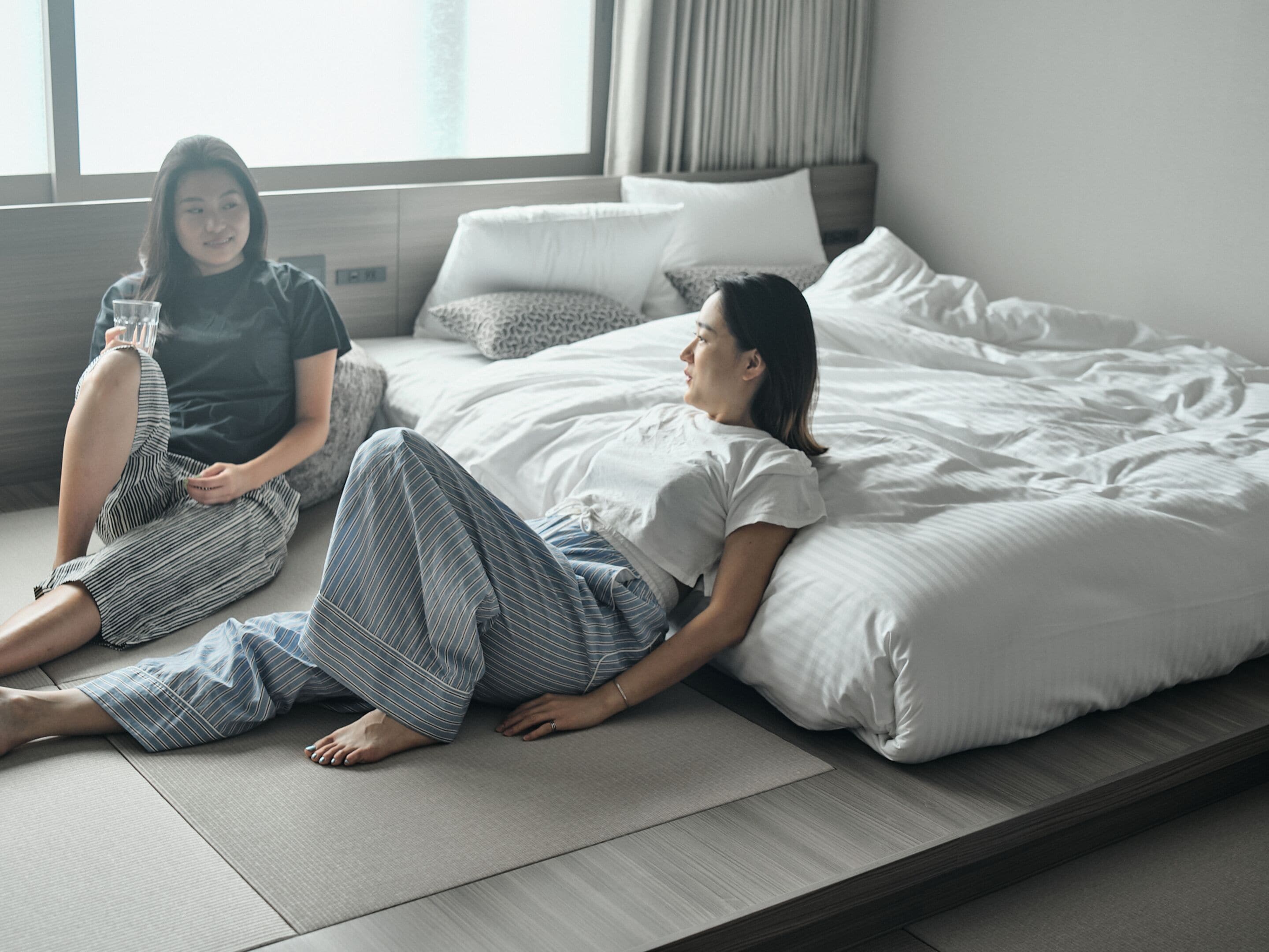 Two people sitting on a bed in a room with a large window, a bedside table, and a headboard.