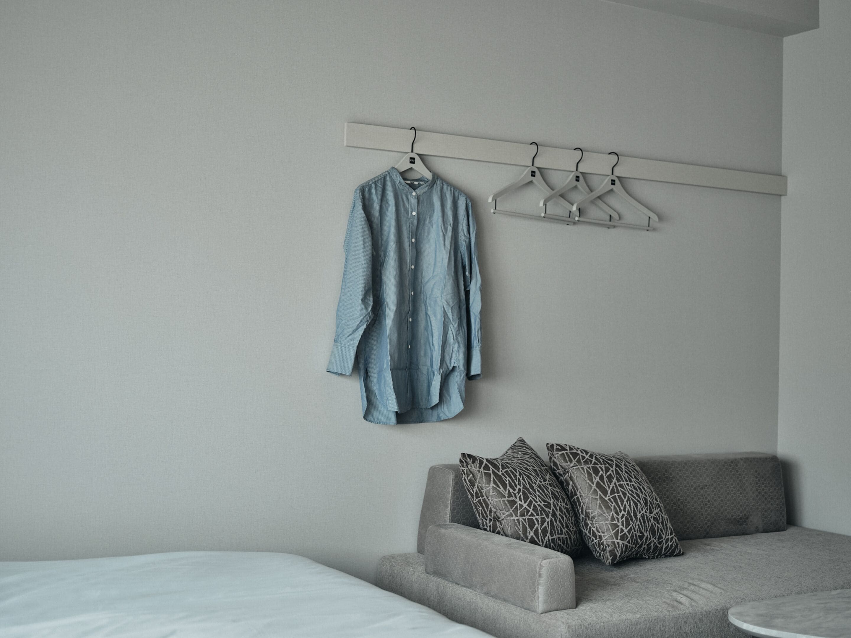 A bedroom with a grey sofa, a bed with white bedding, and a clothing rack with a blue shirt.