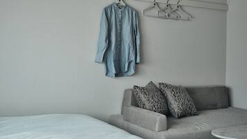 A bedroom with a grey sofa, a bed with white bedding, and a clothing rack with a blue shirt.