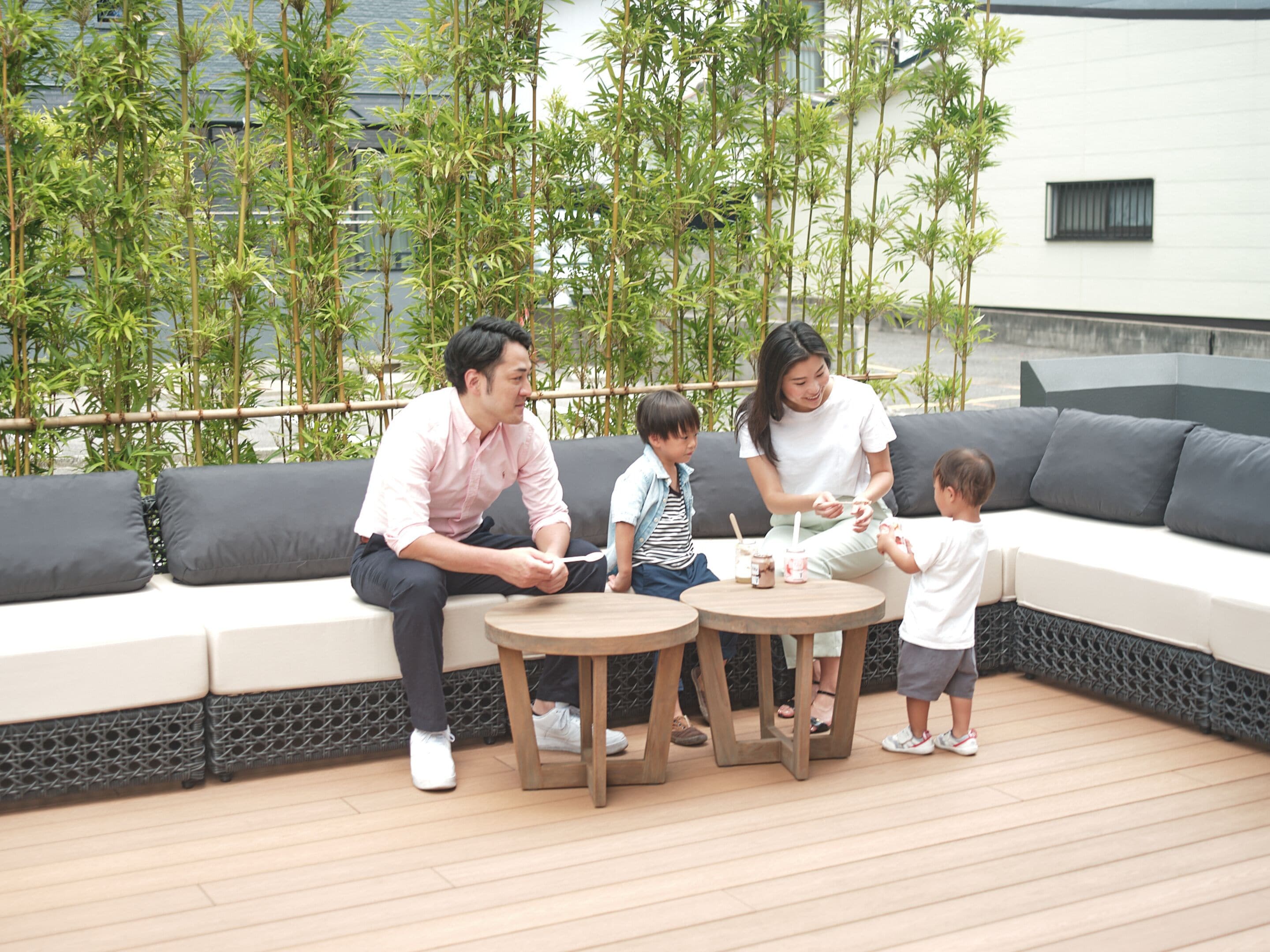 En familie på fire sidder på en moderne udendørs sofa med en træterrasse og bambusplanter i baggrunden.