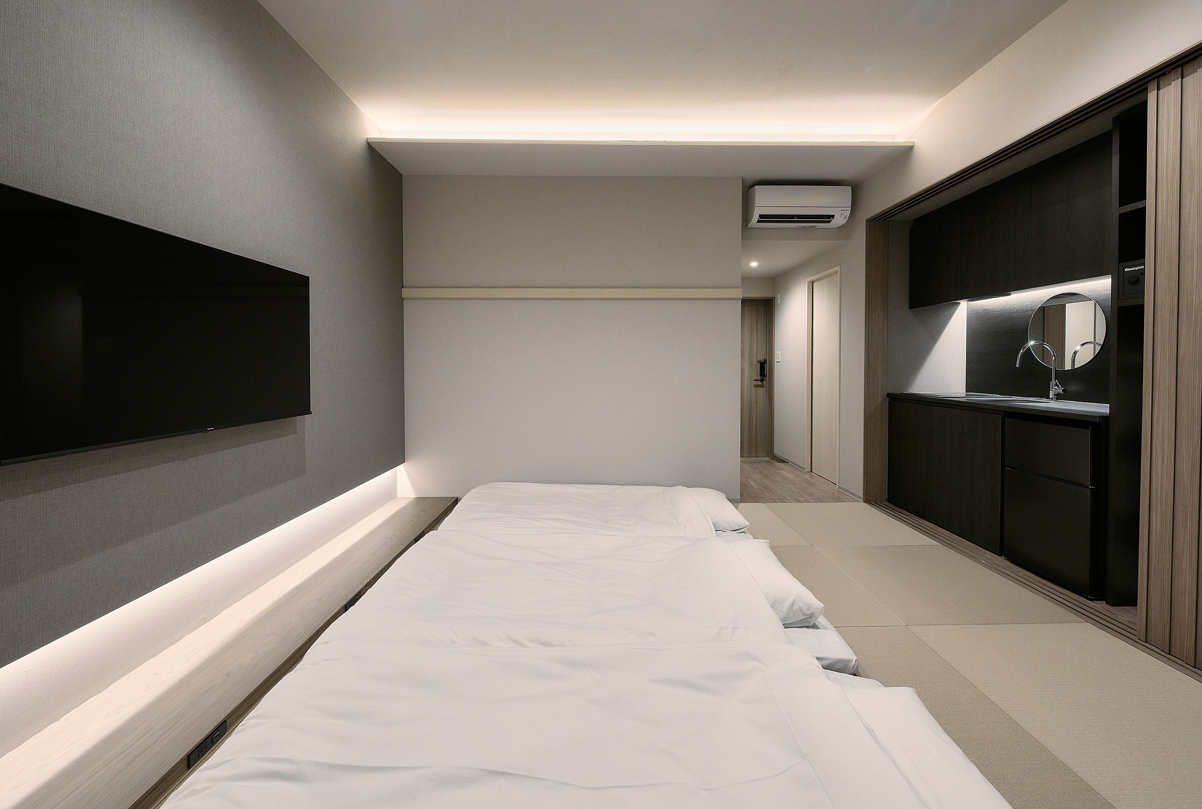 A modern hotel room with a large bed, a TV mounted on the wall, a mirror above the sink, and a walk-in closet.