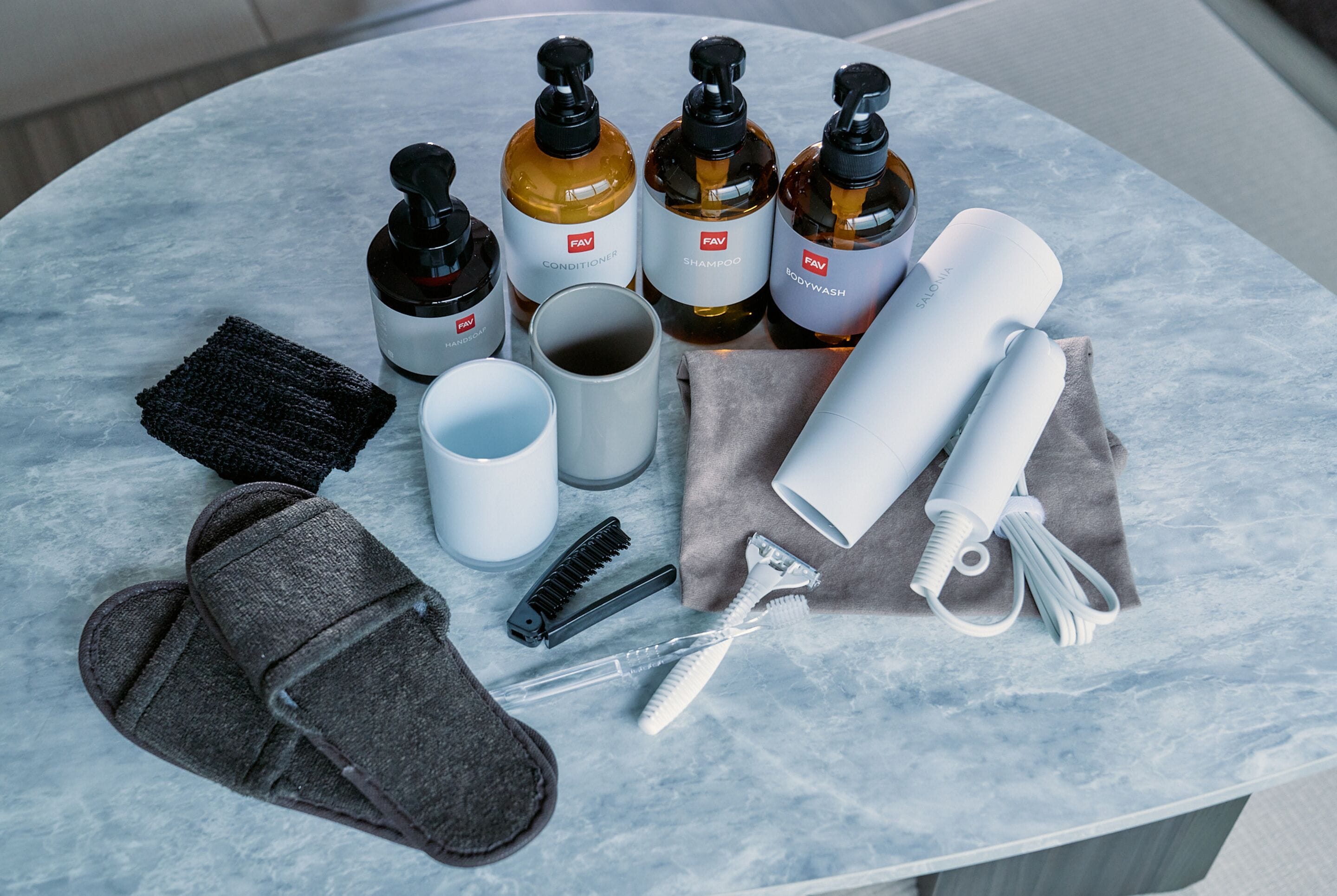 A bathroom vanity with a marble countertop, containing a variety of personal care items including bottles of shampoo, conditioner, and body wash, a razor, a comb, a towel, and slippers.