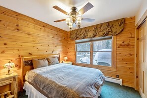A wooden cabin bedroom with a bed, nightstands, a ceiling fan, and a window with a view of the outside. - Cozy Vermont Log Cabin Chalet Only Minutes from Jay Peak (Montgomery)