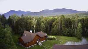 A cabin with a sloped roof, surrounded by dense forest and mountains in the background. - Cozy Vermont Log Cabin Chalet Only Minutes from Jay Peak (Montgomery)