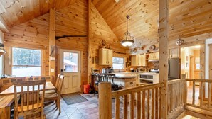 Interior de una cabaña de madera con comedor, cocina y un balcón con barandilla.