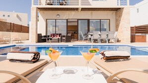 A poolside area with lounge chairs, a pool, and a building with a balcony in the background. - Villa Oleander - 4 bedroom Villa with Private Heated Pool, Hot Tub, Stunning Views (Paphos)
