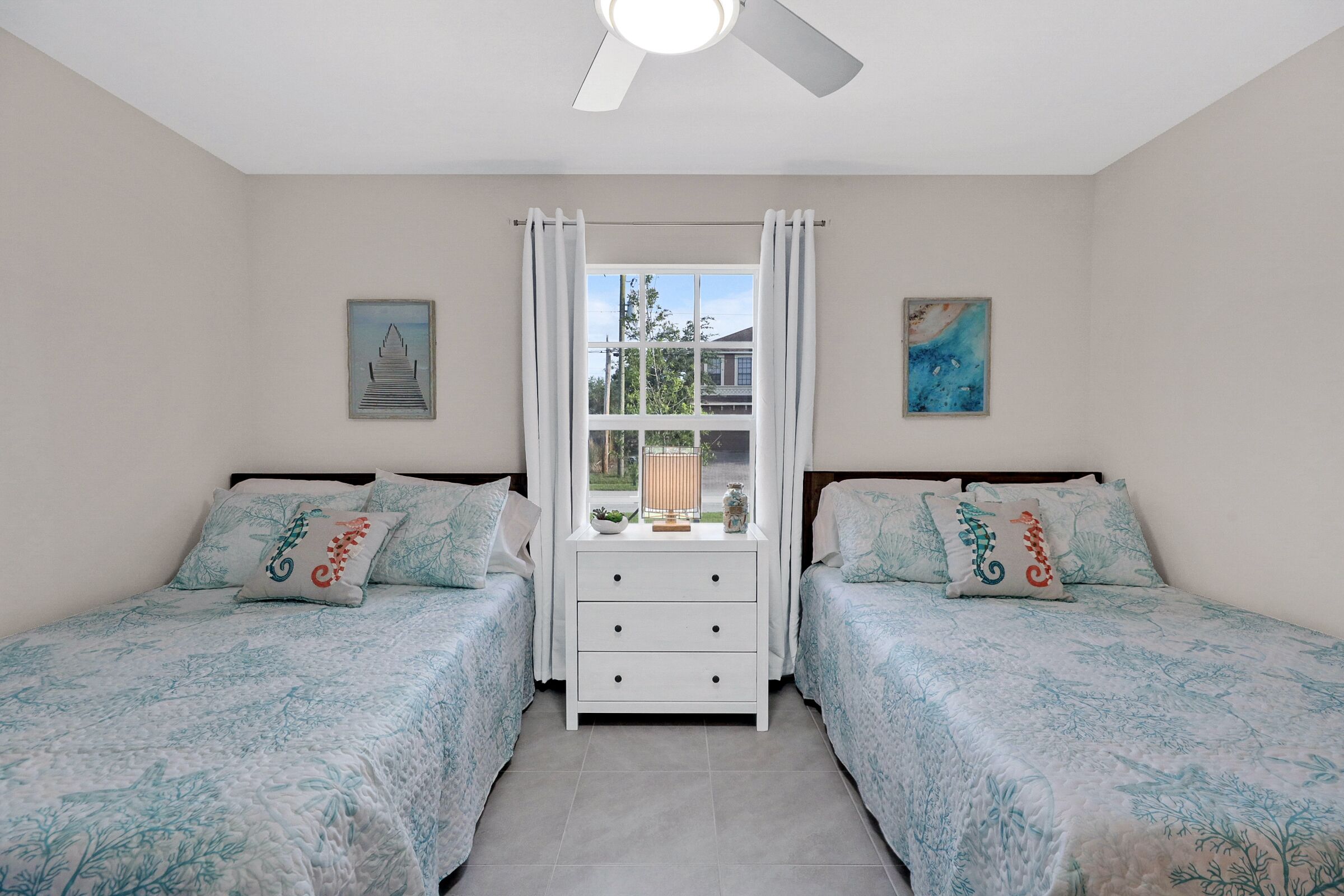 Une chambre avec deux lits, une fenêtre avec des rideaux, un ventilateur de plafond et des œuvres d’art accrochées aux murs.