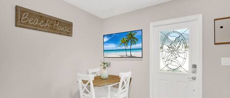 Ein Essbereich mit Holztisch und weißen Stühlen, einem Schild mit der Aufschrift 'Beach House', einem gerahmten Strandbild und einer Tür mit einem Buntglasfenster.