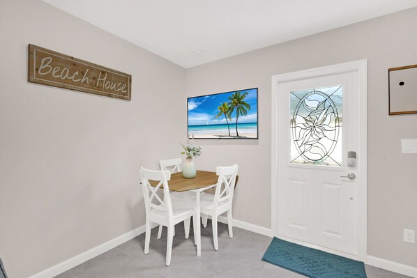 Un comedor con una mesa de madera y sillas blancas, un letrero que dice 'Beach House', una escena de playa enmarcada y una puerta con un vitral.