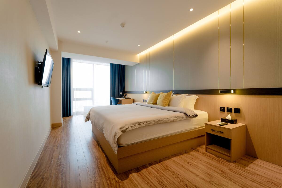Una habitación de hotel moderna con una cama grande, mesitas de noche, un televisor de pantalla plana y un amplio ventanal con cortinas.