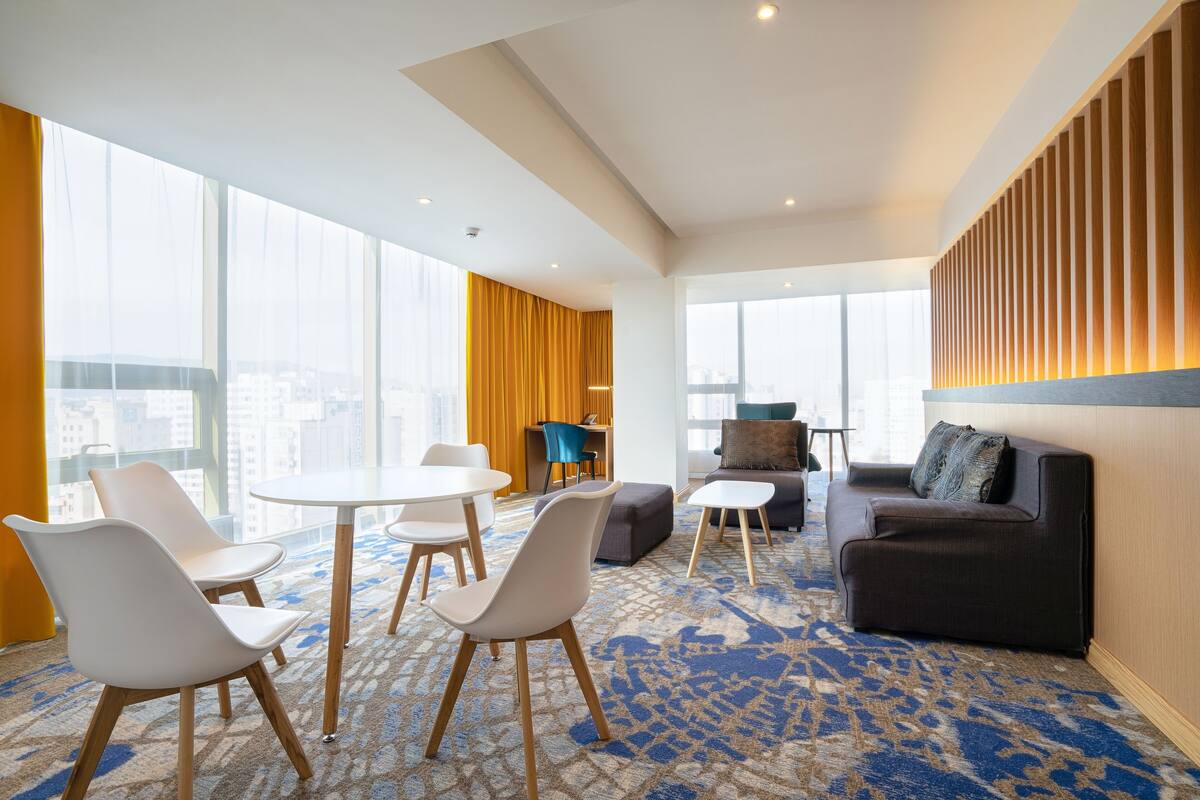 Una habitación de hotel moderna con un ventanal grande, una zona de comedor con sillas blancas, un sofá y una alfombra estampada.