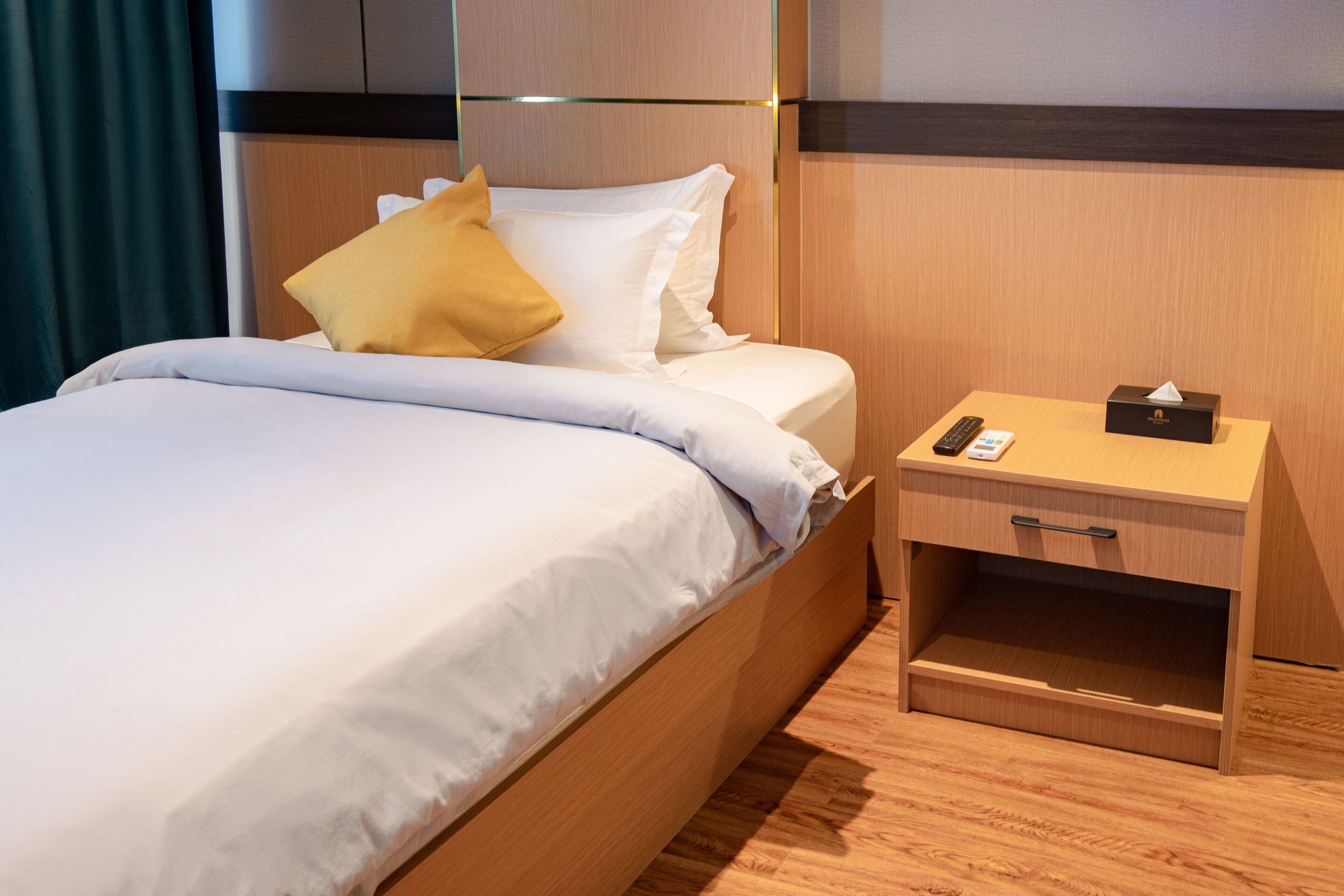 Una cama bien hecha con sábanas blancas y una almohada amarilla, una mesita de noche con un control remoto, una caja de pañuelos y un cajón.