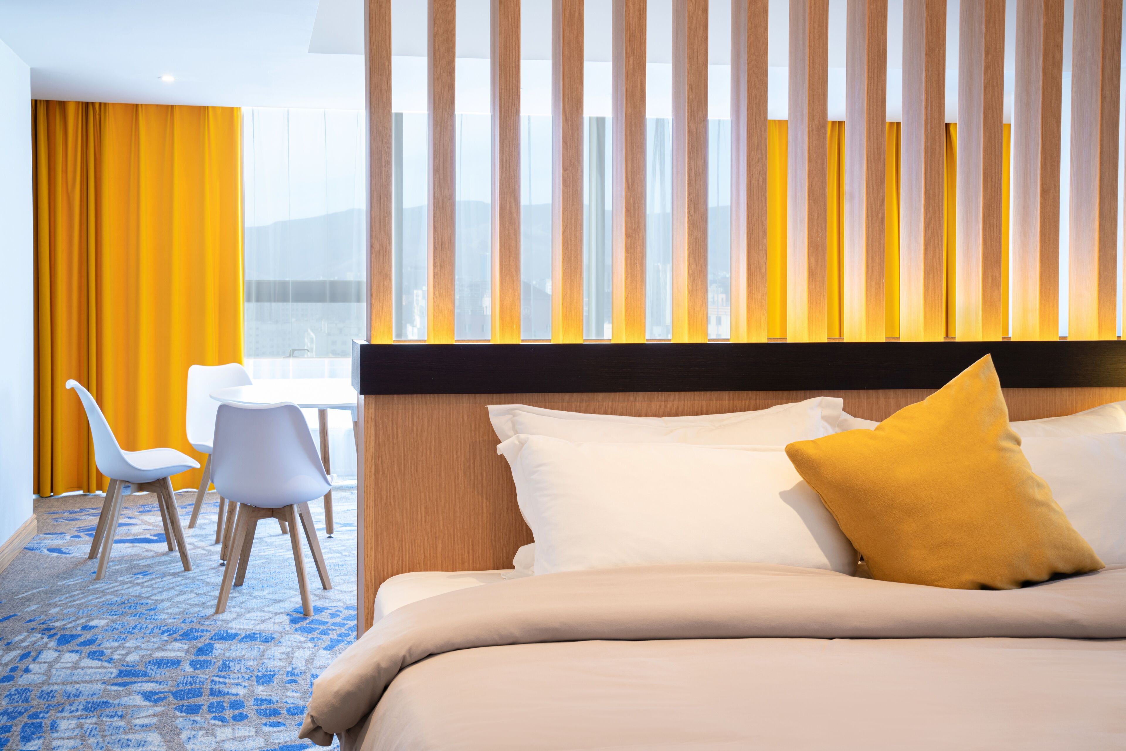 Habitación de hotel con cama, escritorio, dos sillas y vista de la ciudad a través de amplias ventanas.