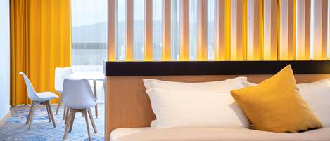 Habitación de hotel con cama, escritorio, dos sillas y vista de la ciudad a través de amplias ventanas.