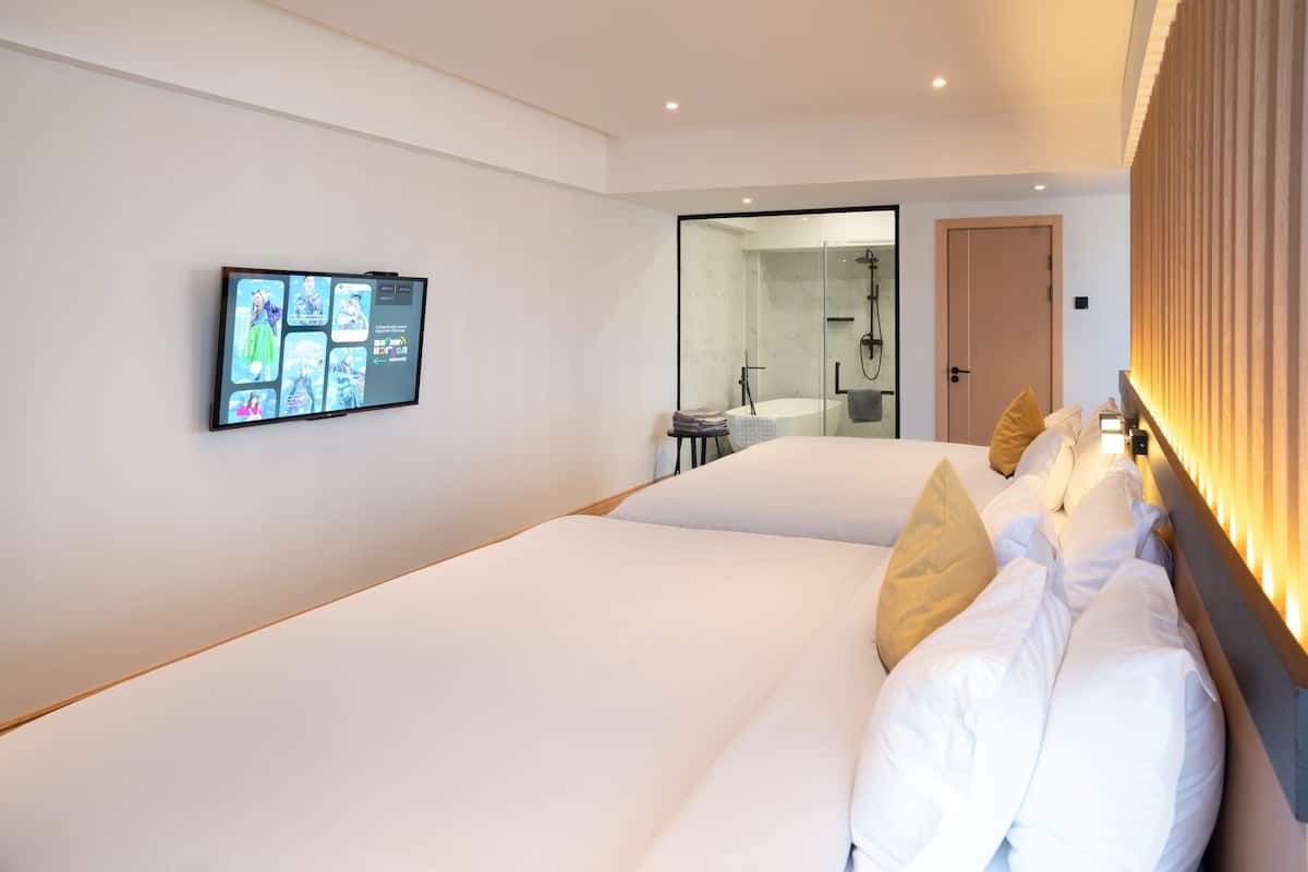 Una habitación de hotel moderna con una cama grande, un televisor montado en la pared y un baño visible a través de una puerta de vidrio.