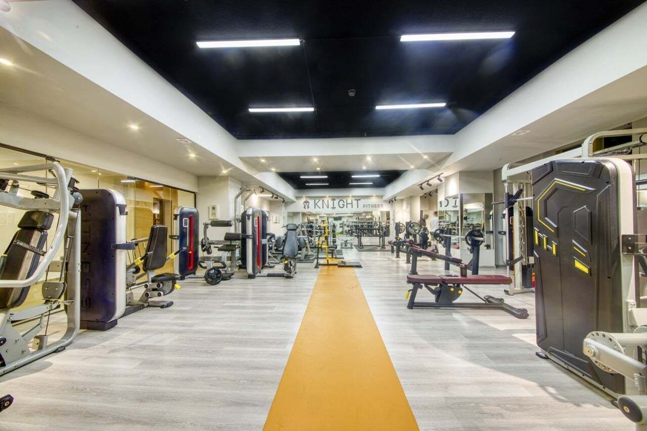 Ein Fitnessstudio mit verschiedenen Trainingsgeräten, einem zentralen Laufweg und einem Schild mit der Aufschrift 'KNIGHT FITNESS'.