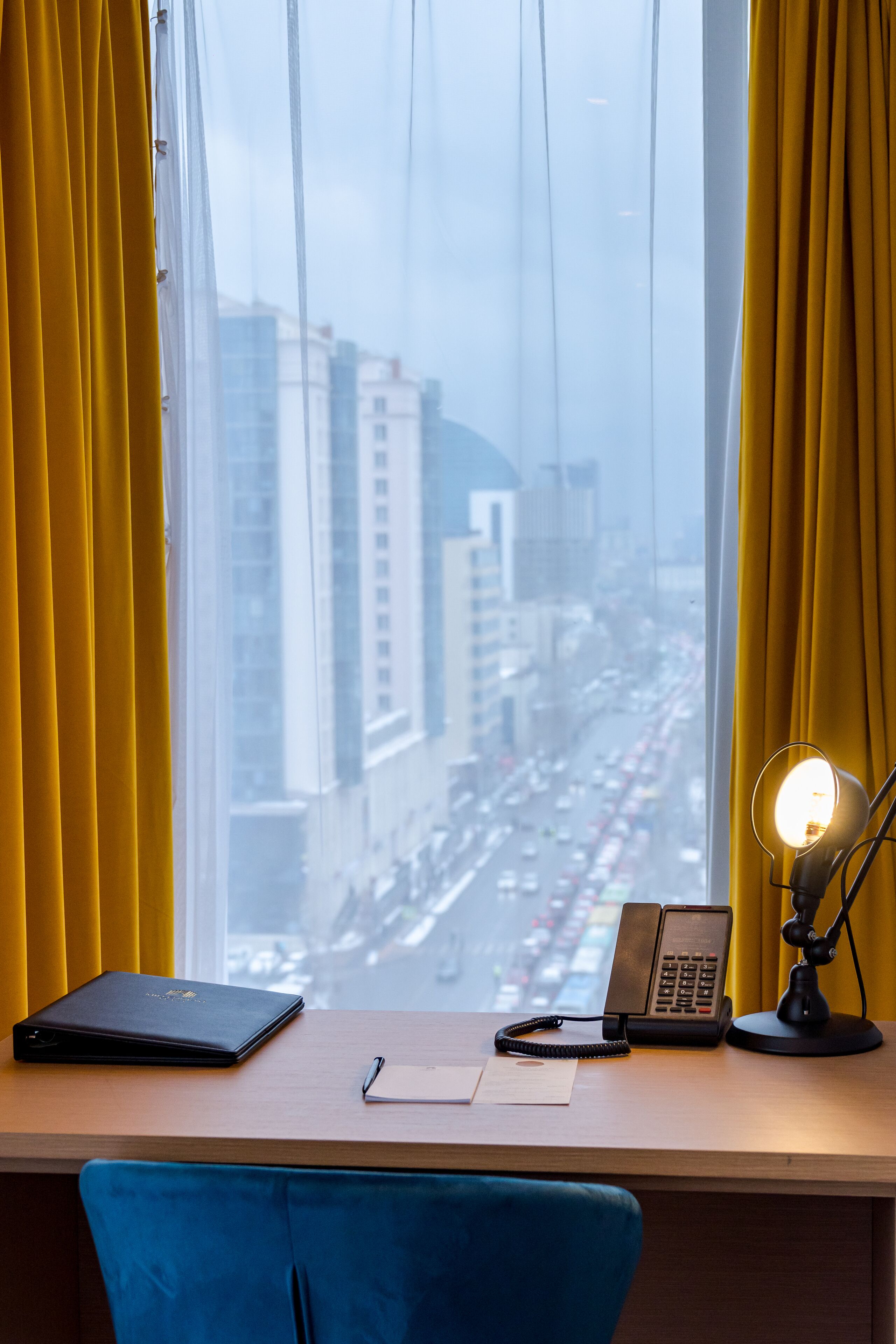 A desk with a laptop, a phone, and a lamp, positioned by a window with yellow curtains, overlooking a cityscape.
