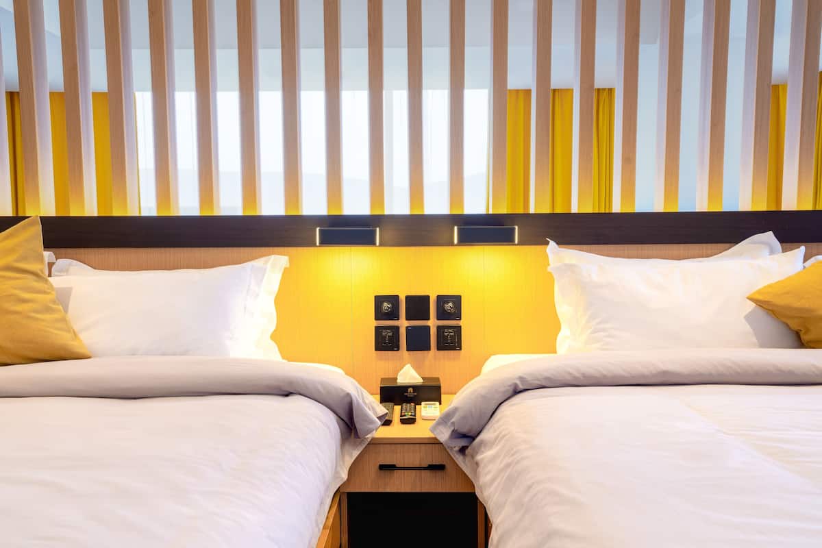 Habitación de hotel con dos camas, cabecera de madera, mesita de noche con lámpara y panel de control en la pared.