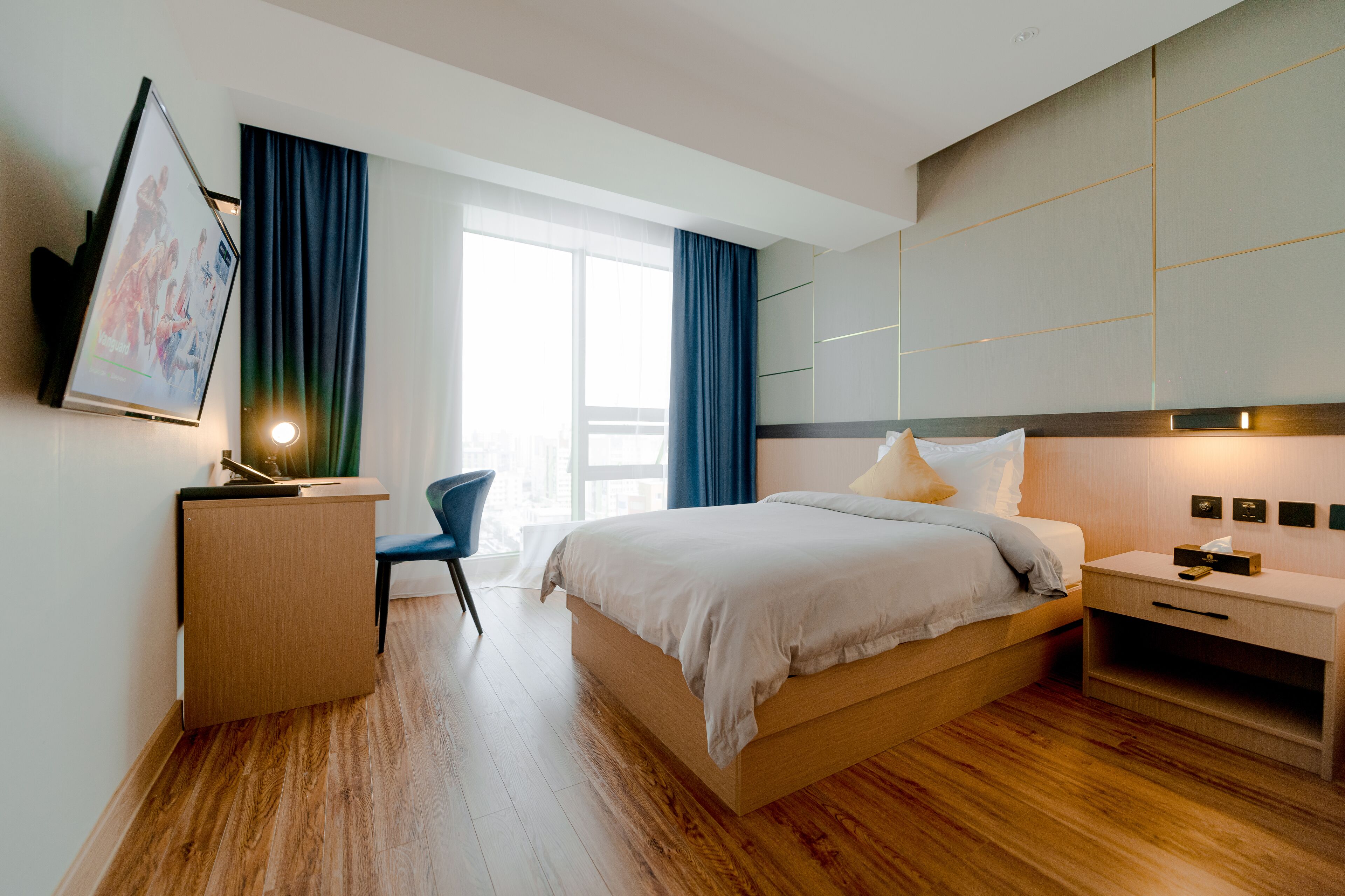 A modern hotel room with a wooden floor, a bed with a white duvet, a desk with a flat-screen TV, a chair, and a bedside table with a lamp.