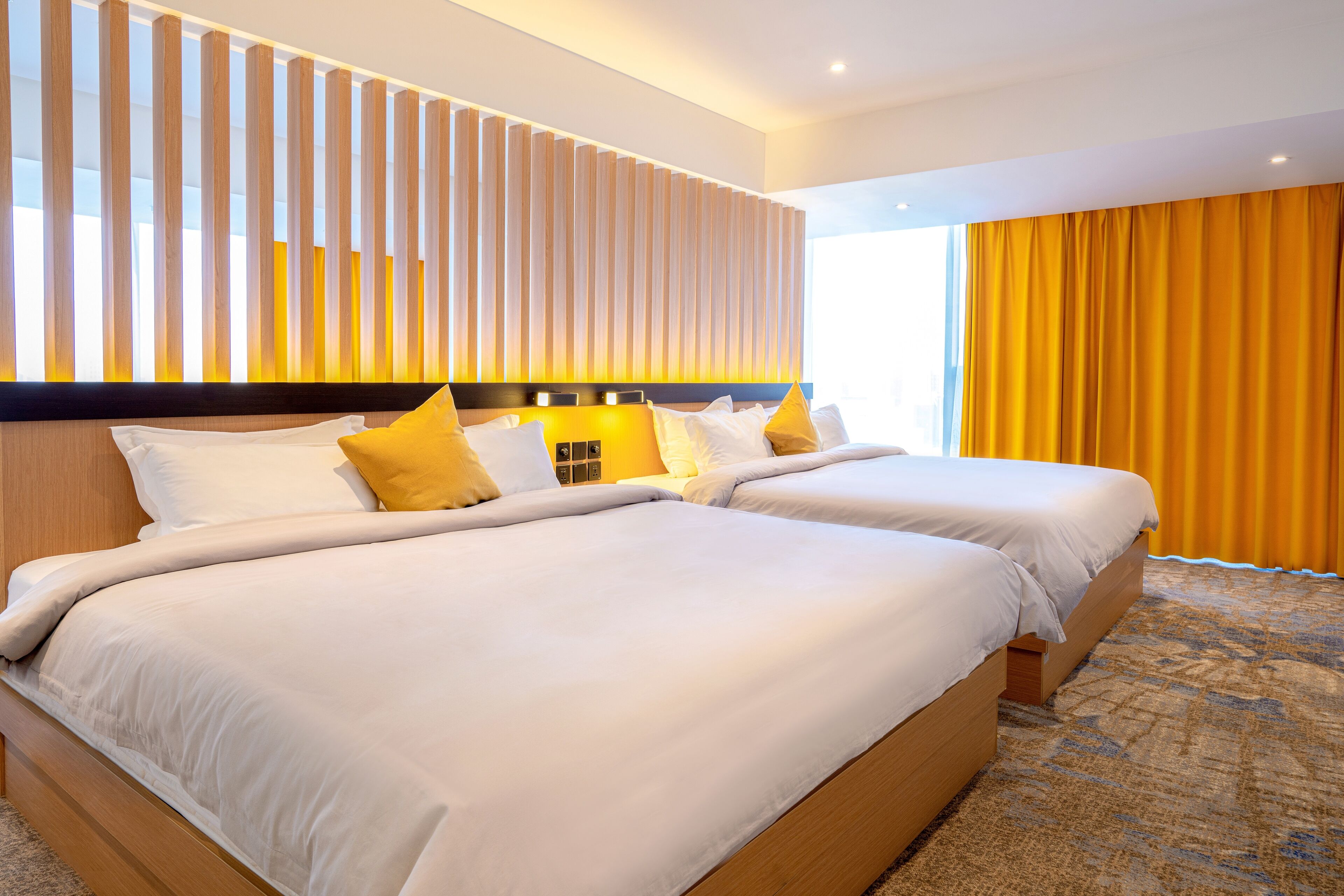 Une chambre d’hôtel avec deux lits, une tête de lit en bois et une grande fenêtre ornée de rideaux jaunes.