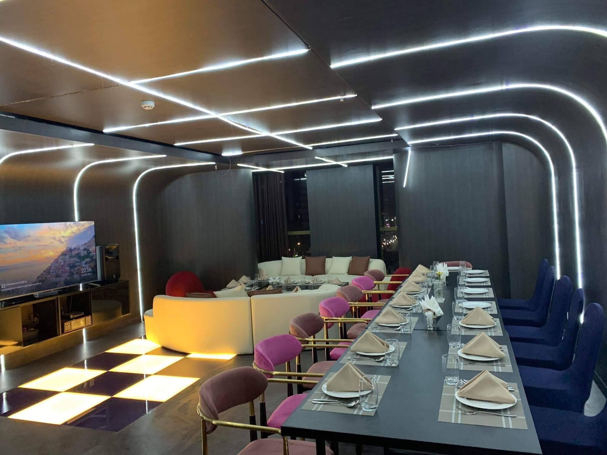 Una sala de conferencias moderna con una larga mesa dispuesta para una reunión, rodeada de asientos cómodos y una pantalla grande para presentaciones.