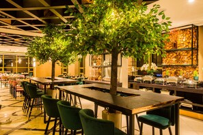 A restaurant with multiple dining tables and chairs, a decorative tree with lights, and a stone wall with shelves. - Millennium Plaza Hotel (Ulaanbaatar)