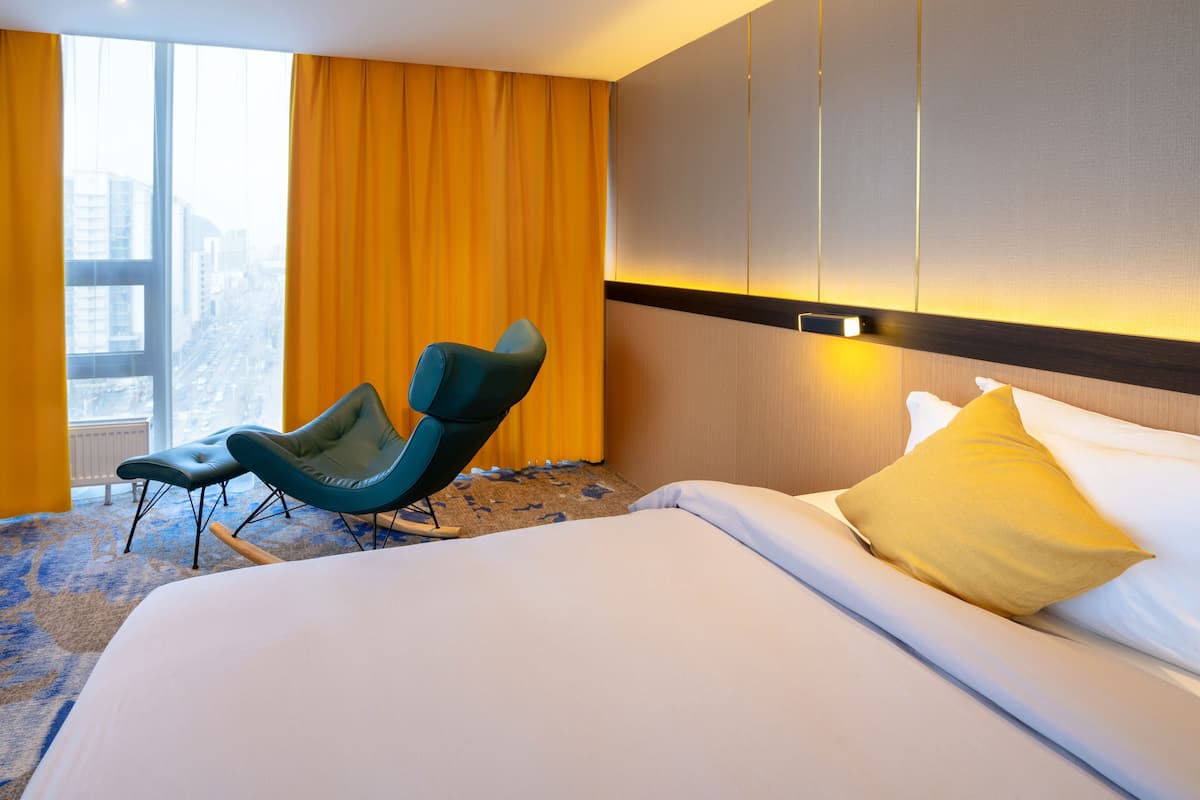 Habitación de hotel con una cama grande, una silla azul, un almohadón amarillo y vistas a la ciudad a través de un ventanal amplio.