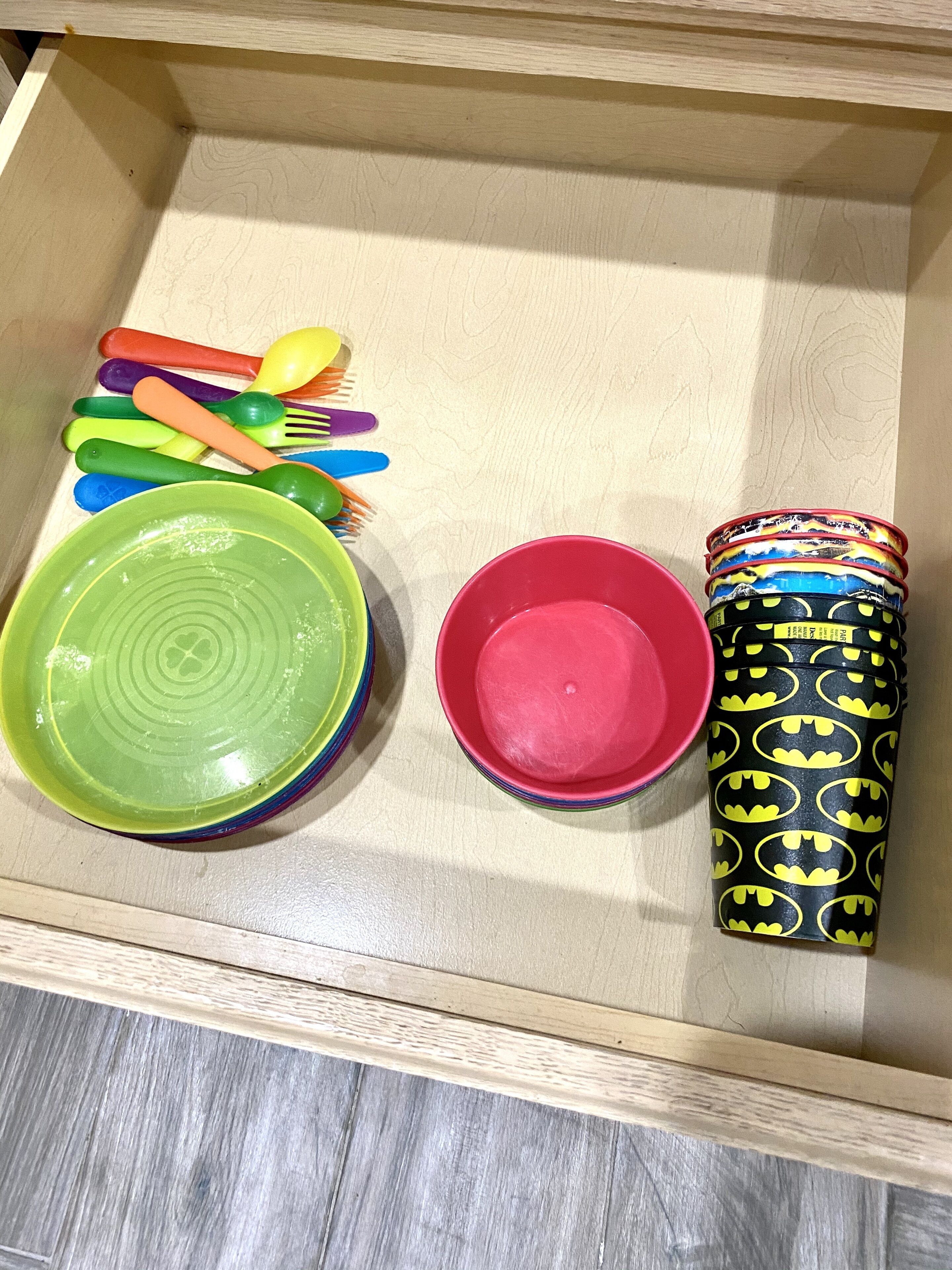 En skuffe med farverige plastbestik, en grøn skål, en lyserød skål og et Batman-tumbler.