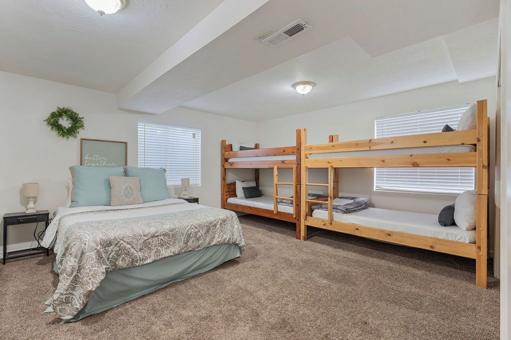 Une chambre avec un grand lit, deux lits superposés, une table de chevet, une lampe fixée au mur et une fenêtre avec des stores.