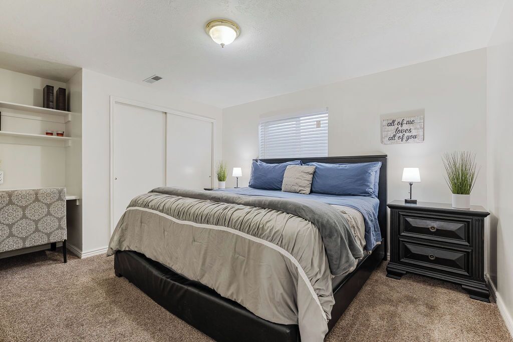 A bedroom with a bed, bedside tables, a nightstand, a window with blinds, a shelf with books, and a wall with a decorative sign.