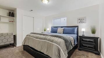 A bedroom with a bed, bedside tables, a nightstand, a window with blinds, a shelf with books, and a wall with a decorative sign.