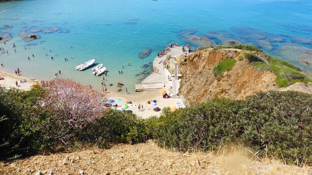 Una playa costera con aguas cristalinas, acantilados rocosos y una zona arenosa con sombrillas y gente.
