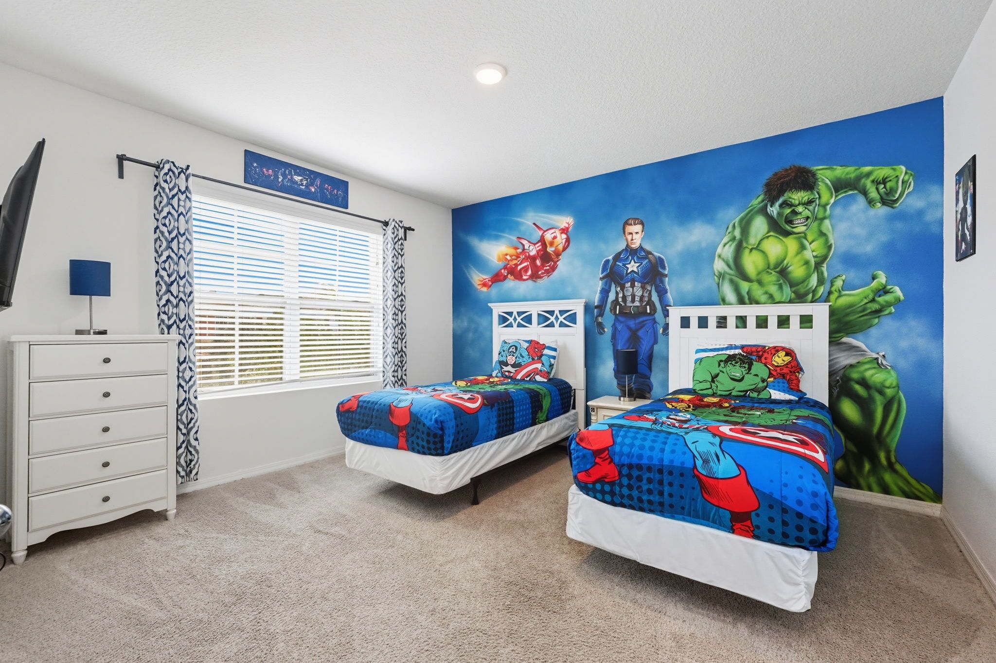 A bedroom with two beds, a dresser, a window with blinds, and a wall mural featuring superheroes.