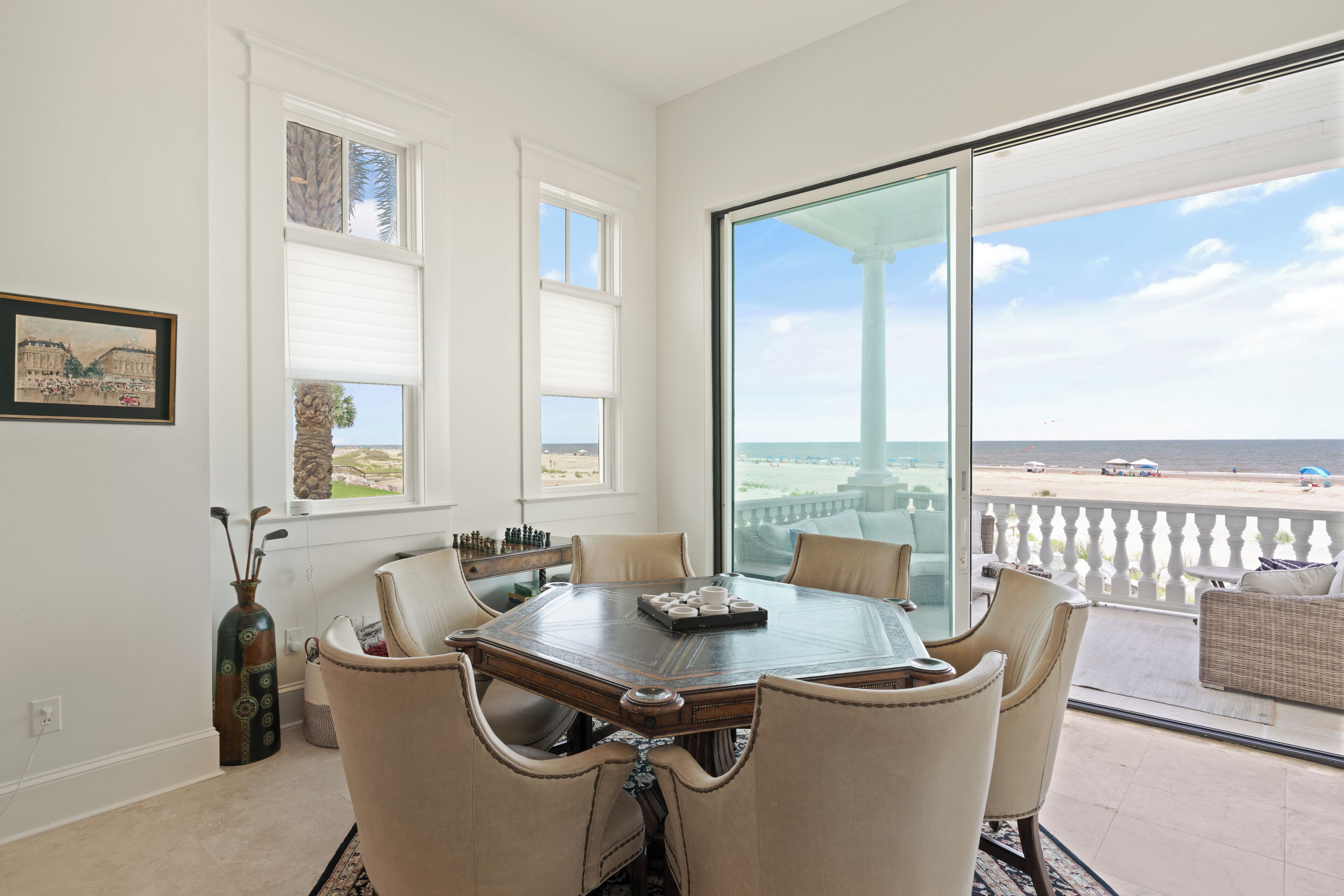 A dining area with a large table and chairs, a vase, and a window with a view of the beach.