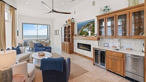 Amplia sala de estar con chimenea, un ventanal grande con vista al mar y una cocina moderna con armarios de madera y electrodomésticos de acero inoxidable.