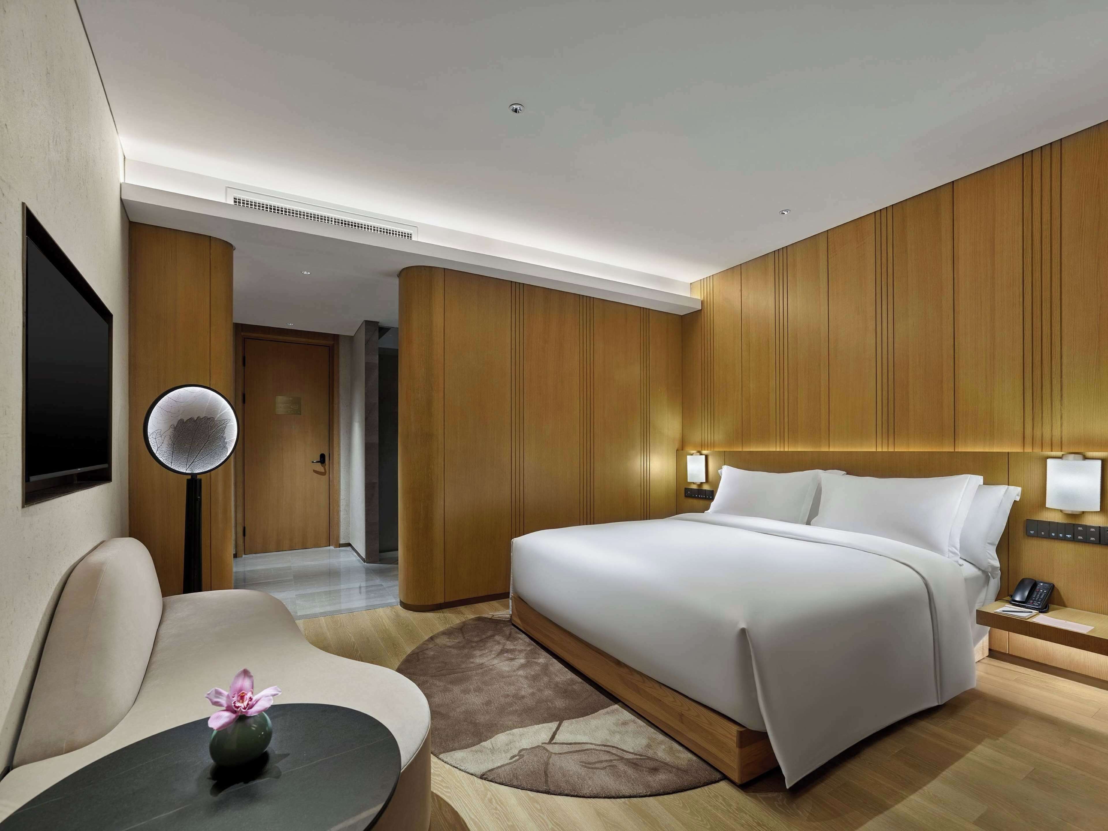 A modern hotel room with a large bed, a desk with a chair, a round table with a vase, and wooden paneling on the walls.