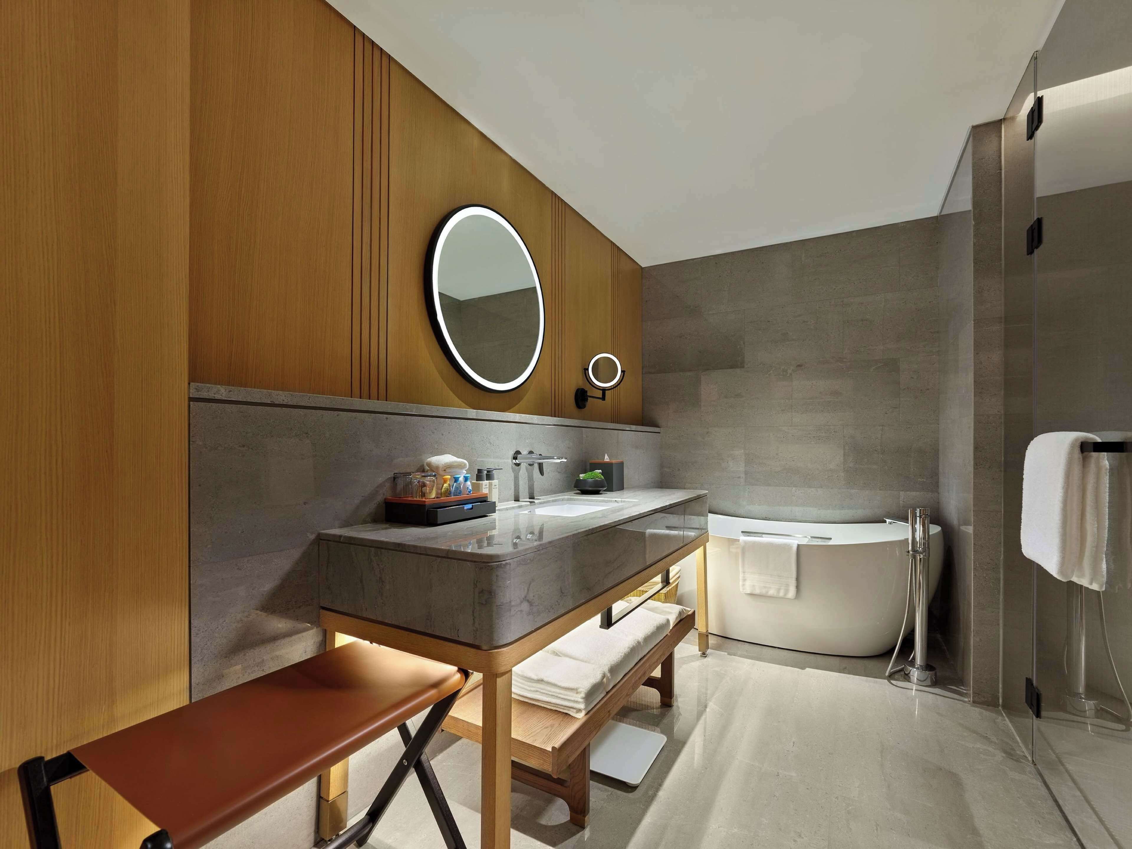 Ein modernes Badezimmer mit Waschtisch aus Holz, einer Bank, einem runden Spiegel, einer Badewanne und einem Handtuchhalter.