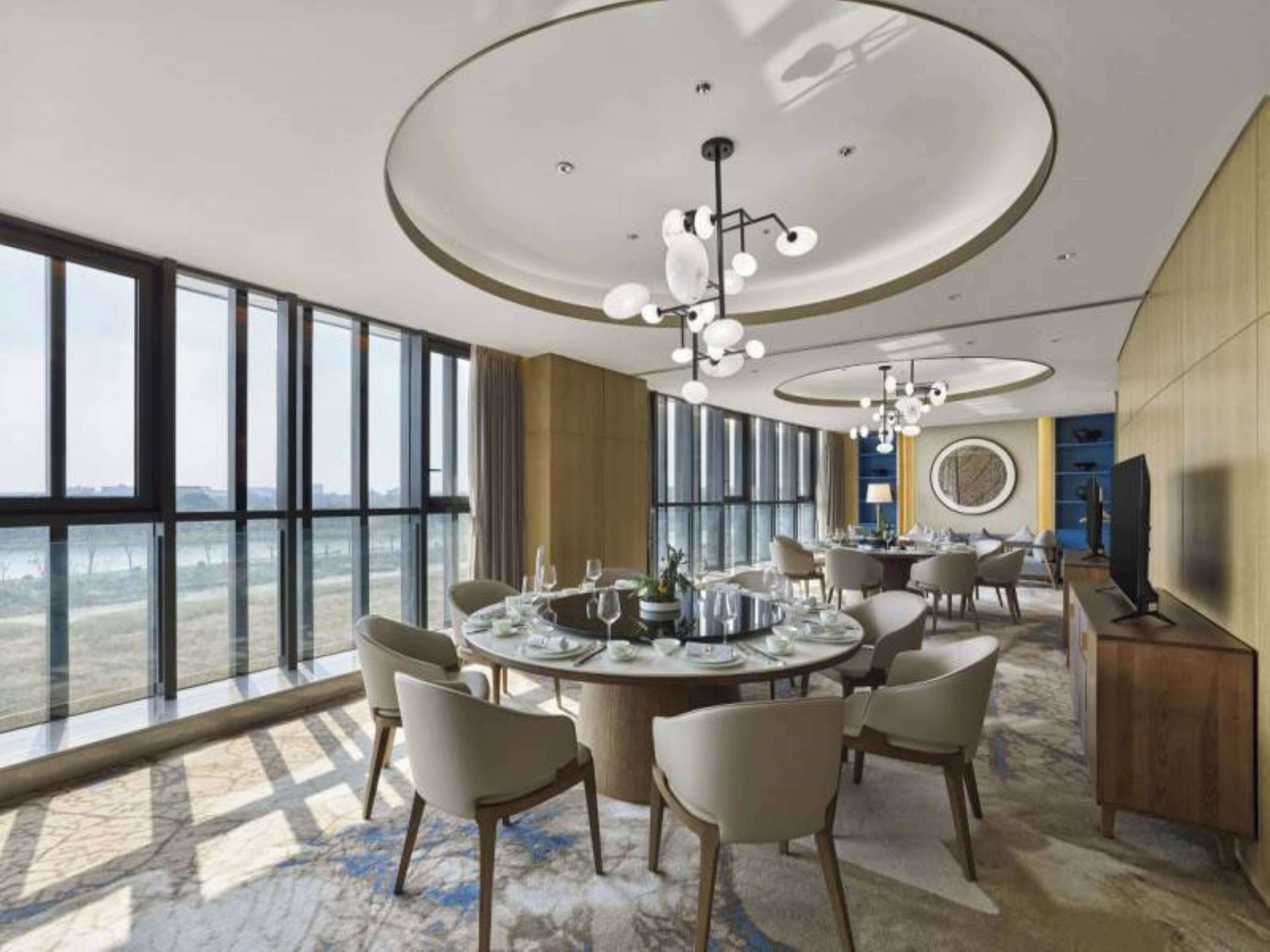 A spacious hotel dining area with large windows, a round table set for dining, and a modern chandelier.