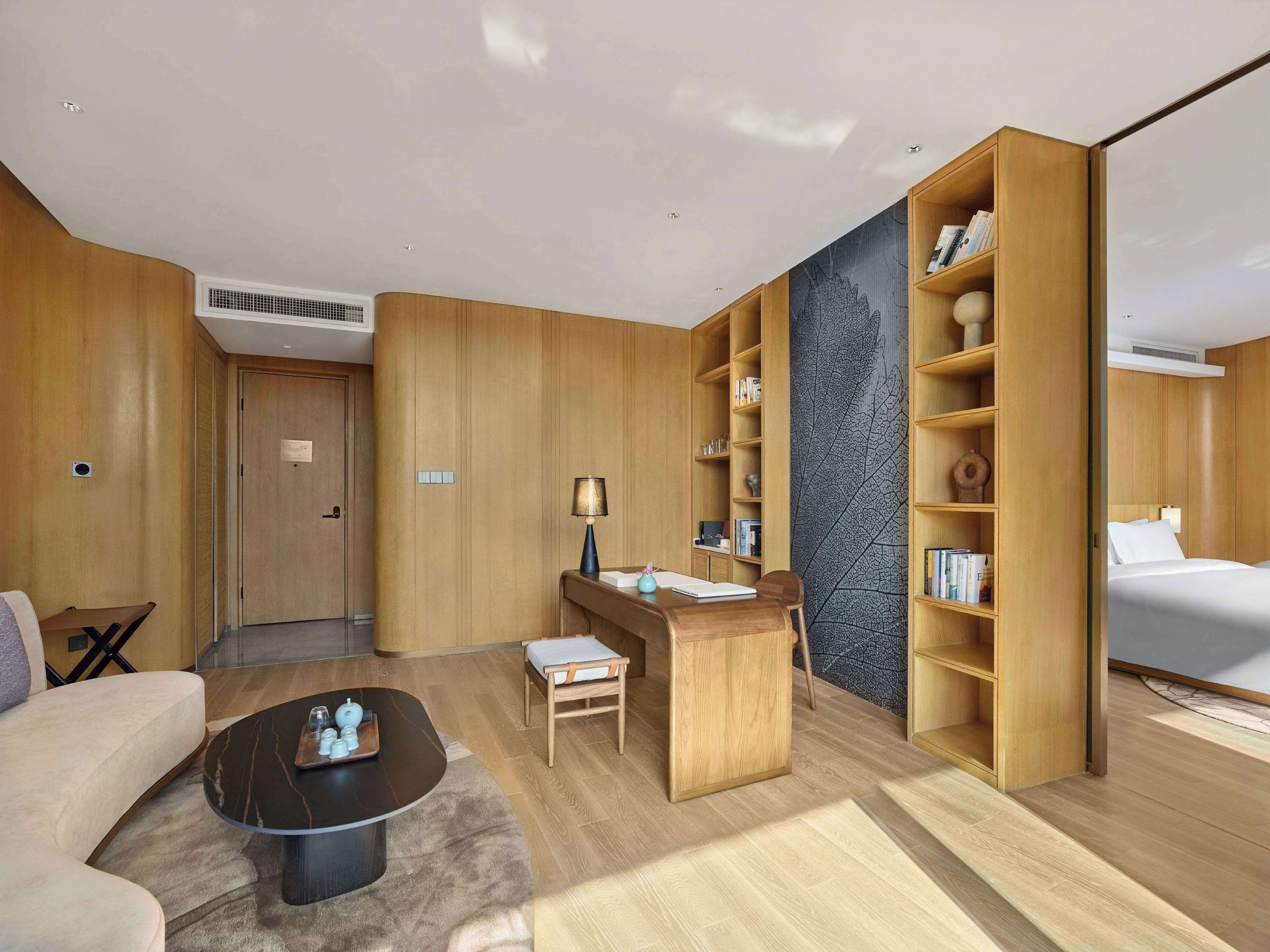 A modern hotel room with a wooden theme, including a desk, bookshelf, and a bed.